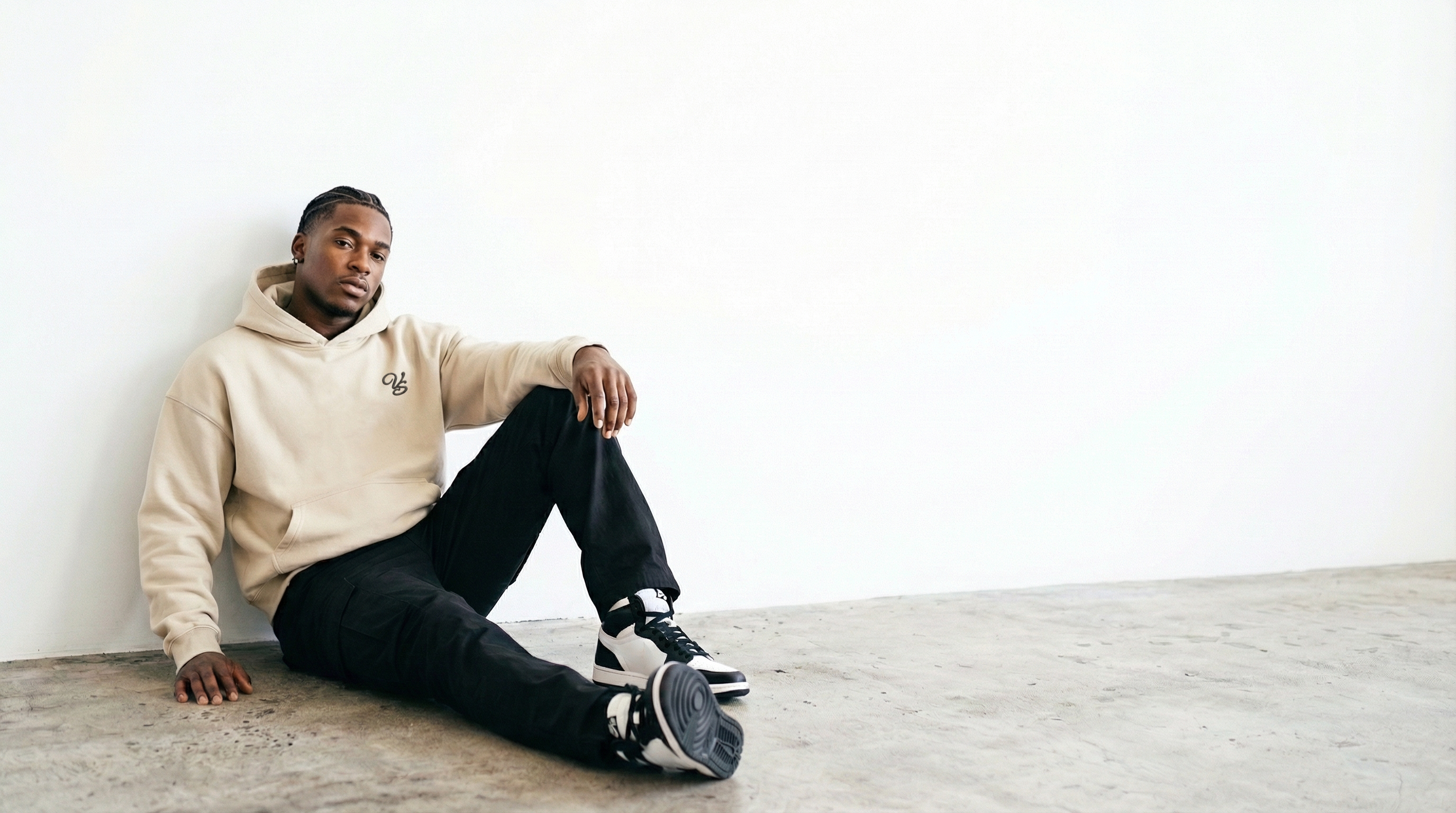 Man sitting on a floor wearing a beige hoodie and black pants in a minimalistic setting.