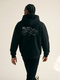Person wearing a black hoodie with mountain graphic and text on the back, standing against a plain background.