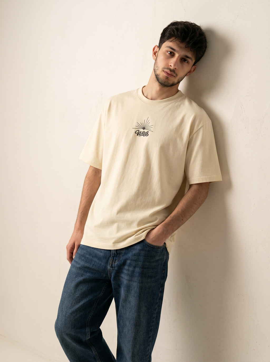 Man wearing a beige t-shirt and blue jeans against a plain background