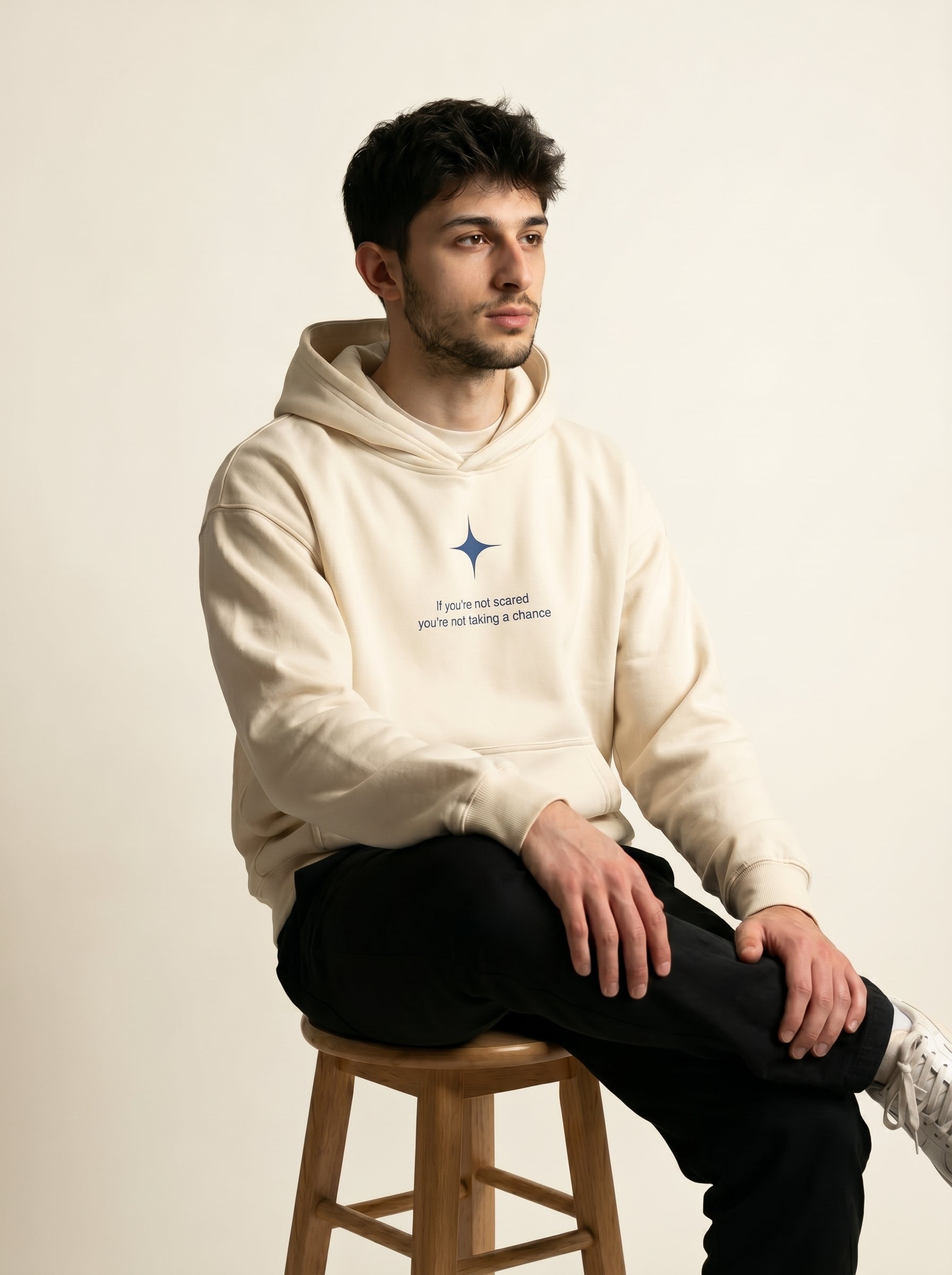 Man wearing a beige hoodie with text and a logo, sitting on a wooden stool against a plain background