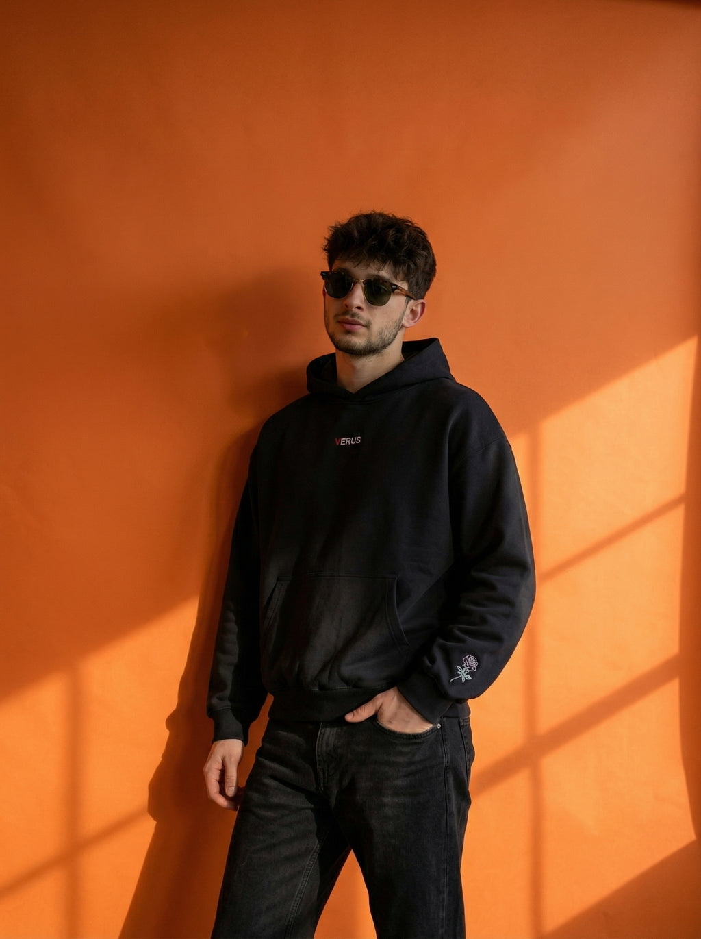Man wearing a black hoodie and sunglasses against an orange wall